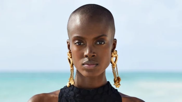 Madisin Rian poses in front of the ocean in a black onesie swimsuit and oversized gold earrings.